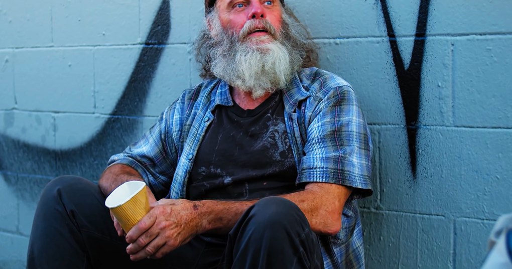 Older adult male wearing plaid short-sleeved shirt, dark t-shirt, black pants, and beanie hat, holding disposable coffee cup in an urban setting with graffiti on a blue brick wall during daytime.