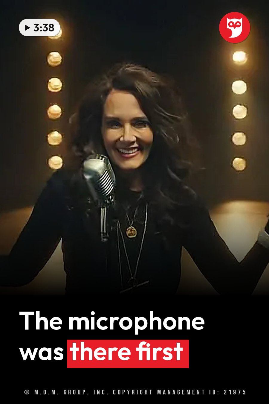 A woman with dark hair smiles while holding a vintage microphone on stage under bright lights. A headline runs in white letters across the lower third.