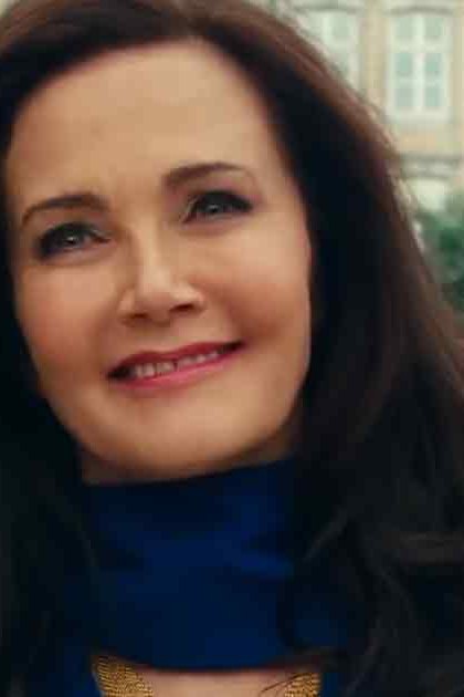 An adult woman wearing a blue turtleneck sweater and black jacket or coat stands at an outdoor market with clear skies, likely spring or summer.