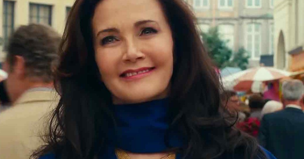 An adult woman wearing a blue turtleneck sweater and black jacket or coat stands at an outdoor market with clear skies, likely spring or summer.