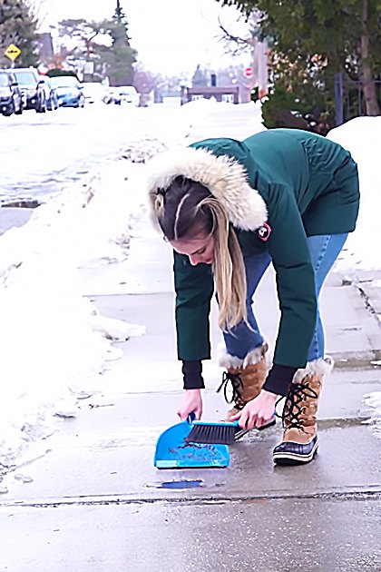 A young adult female wearing a green jacket with fur-lined hood stands outdoors in an urban residential area during winter, surrounded by snow-covered trees and parked cars.