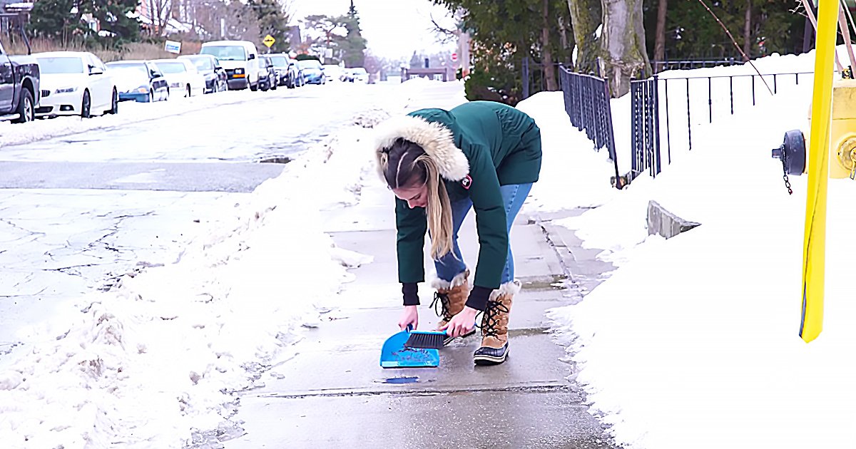 A young adult female wearing a green jacket with fur-lined hood stands outdoors in an urban residential area during winter, surrounded by snow-covered trees and parked cars.