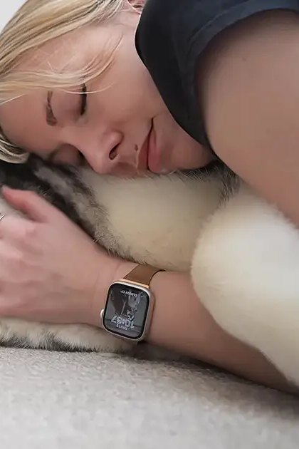An adult female with blonde hair wearing a dark sleeveless top, rings, and smartwatch lies down next to her husky dog in an indoor setting with neutral-colored walls and carpeting. The scene is softly lit, suggesting daytime or early evening.