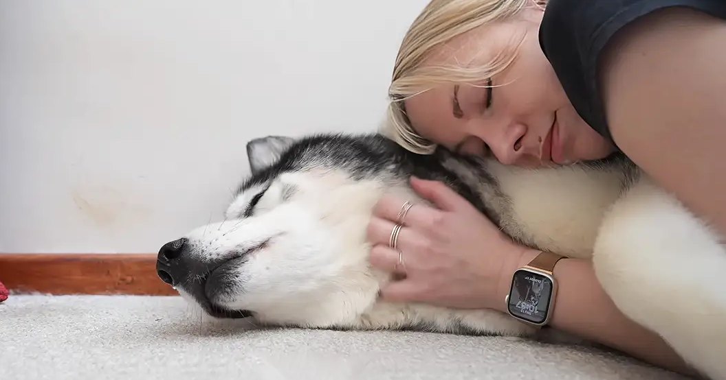 An adult female with blonde hair wearing a dark sleeveless top, rings, and smartwatch lies down next to her husky dog in an indoor setting with neutral-colored walls and carpeting. The scene is softly lit, suggesting daytime or early evening.