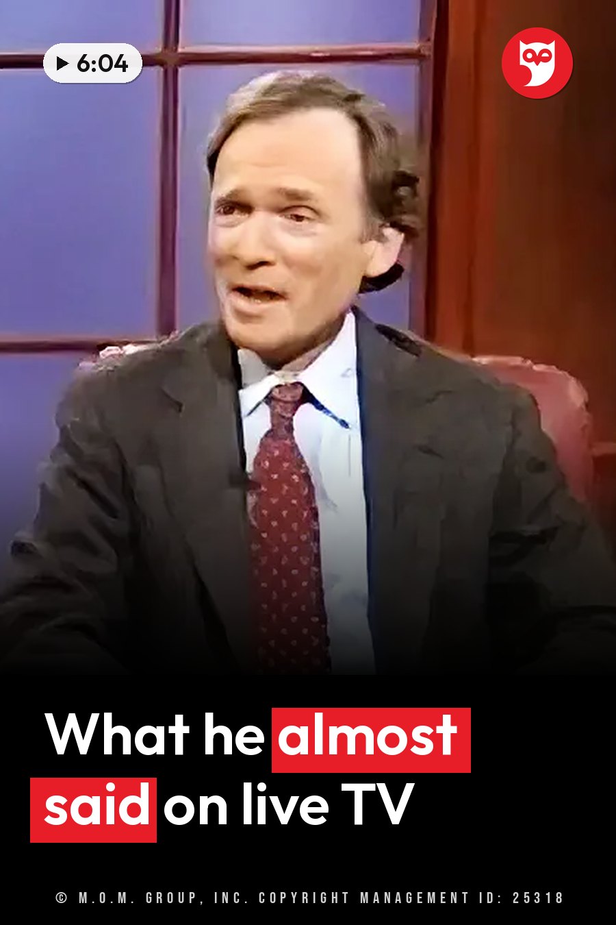 A man in a suit and tie sits in a chair and speaks, his mouth open, in front of a blue paneled wall. A headline runs in white letters across the lower third.