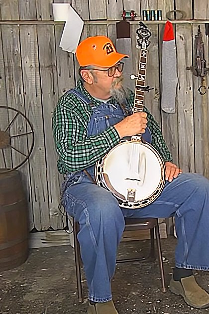 Two older adults playing musical instruments in a rustic old cabin or shed setting.