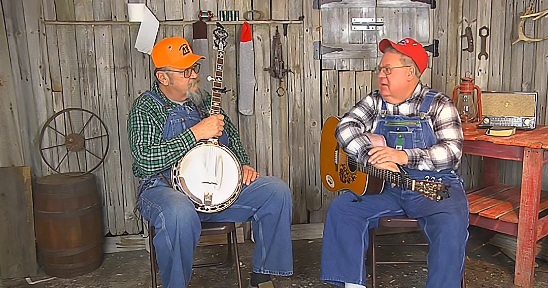 Two older adults playing musical instruments in a rustic old cabin or shed setting.