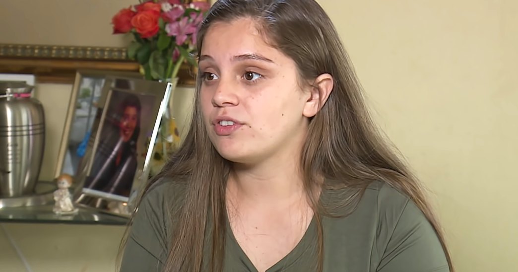 A young adult female with long brown hair, wearing a green V-neck T-shirt indoors. A shelf holds silver urns, a lit candle, framed photograph, and bouquet of red roses in a home setting.