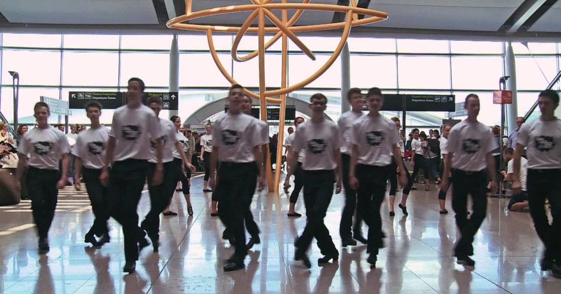 Young adults performing synchronized dance steps in a modern airport terminal with golden sculpture and directional signs visible.
