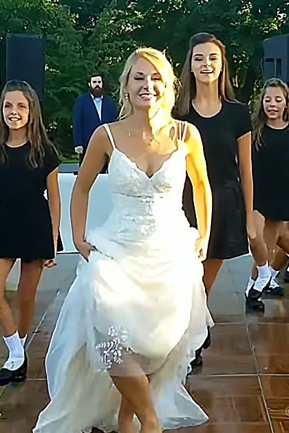 An outdoor wedding reception scene with a bride in a white wedding dress and young adults wearing black dresses, surrounded by greenery and natural light.