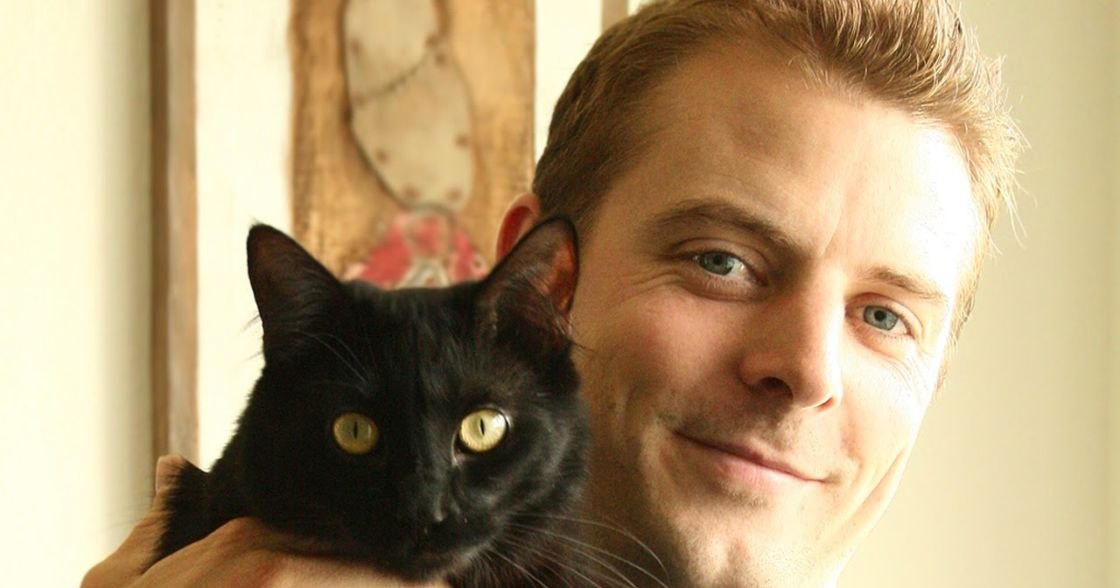 An adult male holding a black cat with yellow eyes indoors, soft natural lighting creates a warm atmosphere