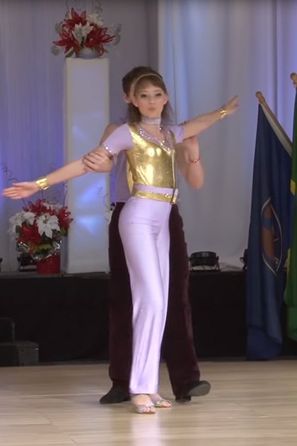 A young adult female dancer wearing a gold leotard and white pants, performing at an 'US Open' dance competition with flags from various countries in the background.