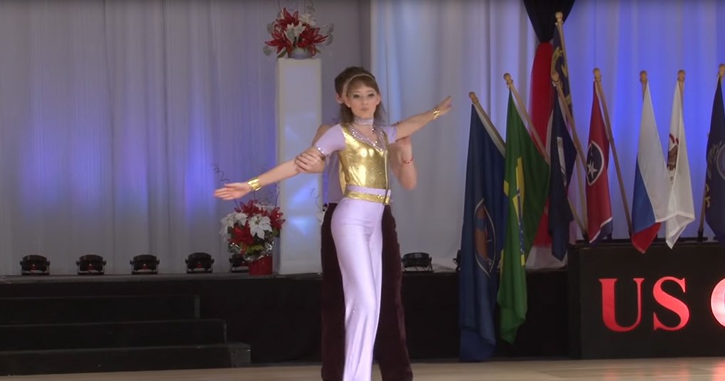 A young adult female dancer wearing a gold leotard and white pants, performing at an 'US Open' dance competition with flags from various countries in the background.