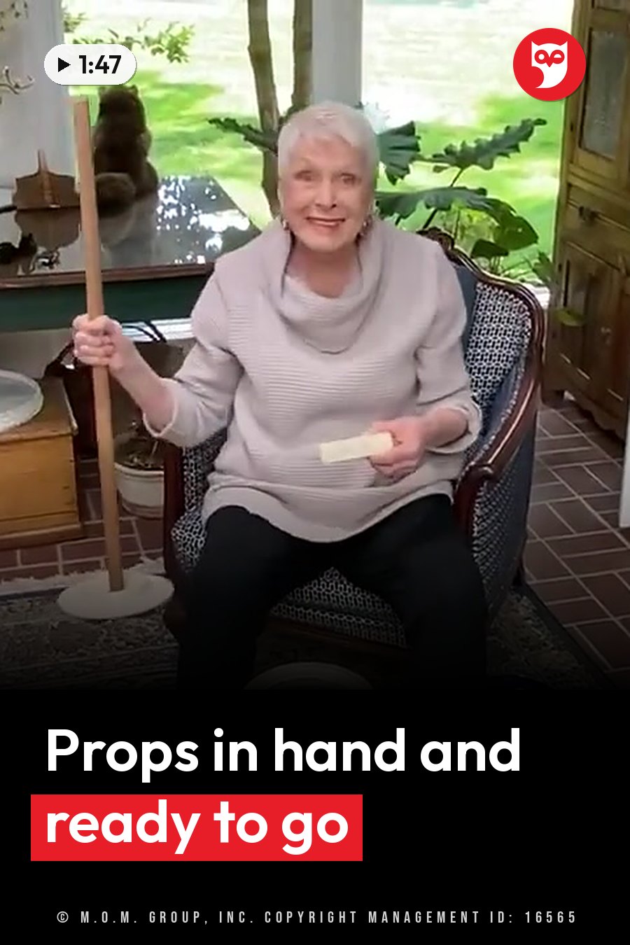 An older woman sits holding a long stick and a small object, smiling at the camera. A headline runs in white letters across the lower third.