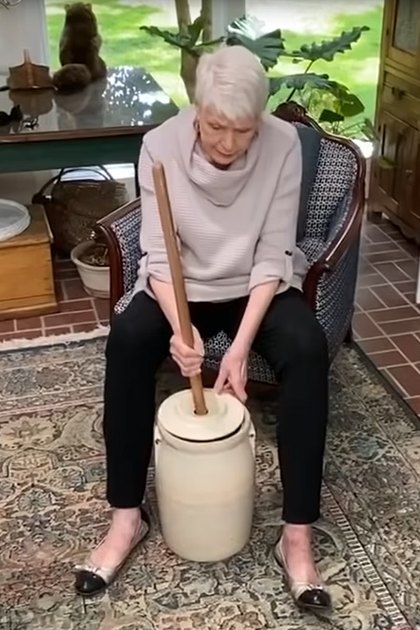 An older adult female wearing a cream sweater with black pants and white shoes in a home setting with greenery visible through windows indicating daytime.