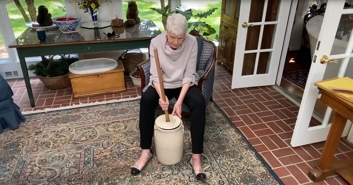 An older adult female wearing a cream sweater with black pants and white shoes in a home setting with greenery visible through windows indicating daytime.