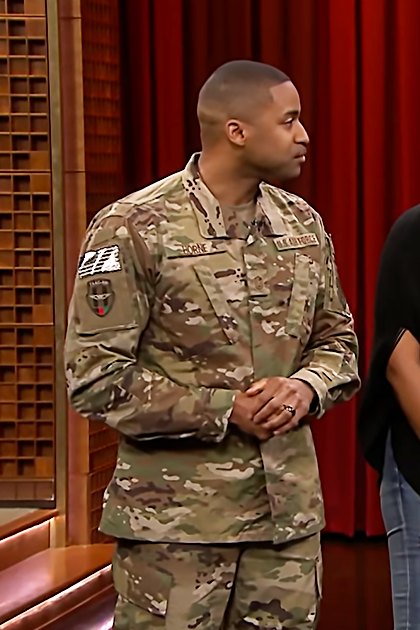 A talk show host in a studio with red curtains, wooden paneling, and a logo reading 'Madly Odd'.
