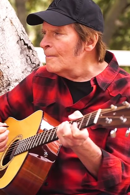 Middle-aged man playing guitar outdoors amidst greenery and a white wooden fence, likely spring or summer.