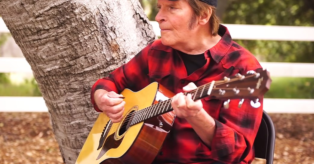 Middle-aged man playing guitar outdoors amidst greenery and a white wooden fence, likely spring or summer.