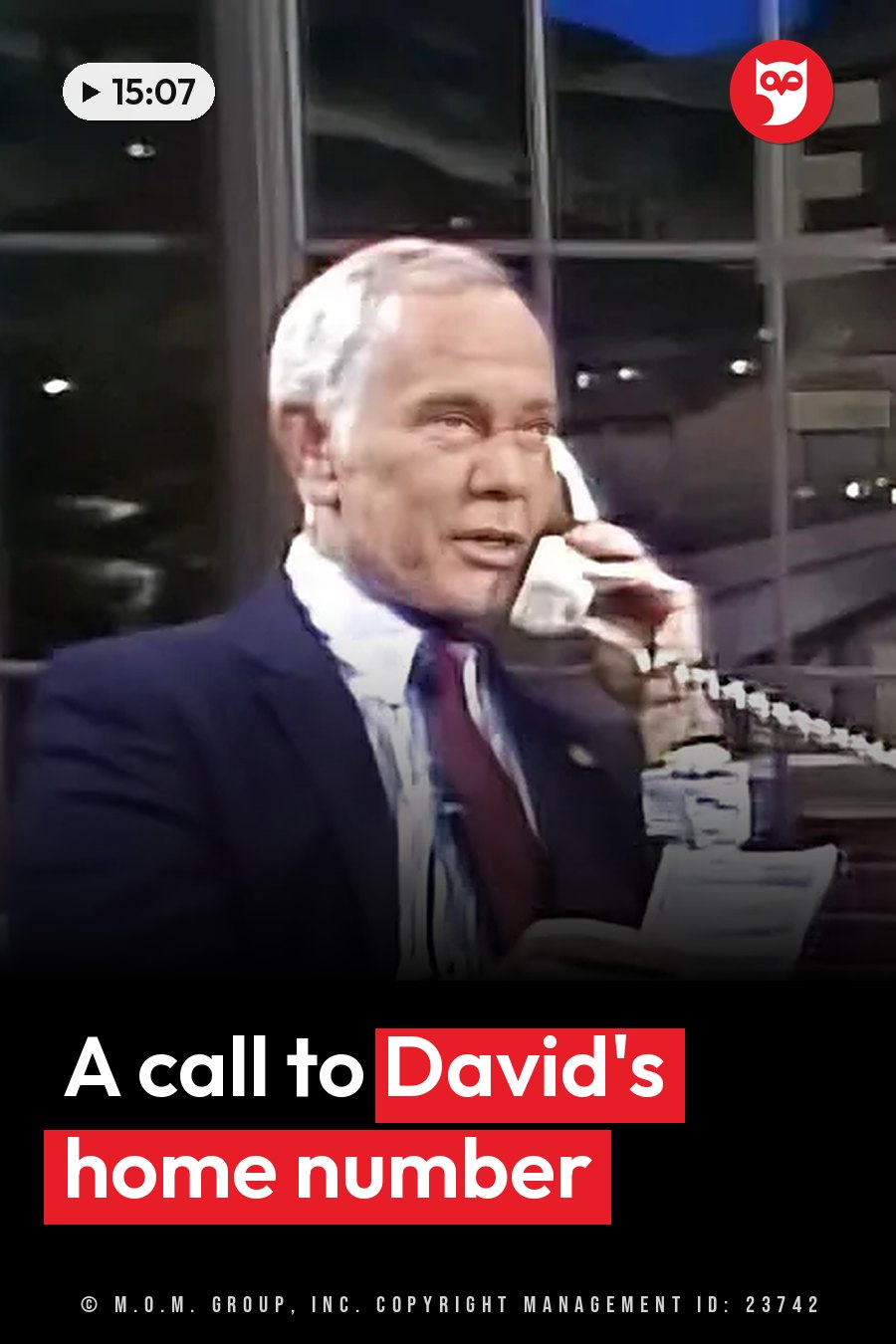 A man in a suit and tie holds a telephone to his ear while seated at a desk with papers nearby. A headline runs in white letters across the lower third.