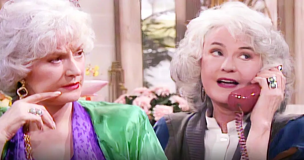 Two older adults dressed elegantly, one wearing a purple garment with green shawl and the other in a dark blazer over a white shirt, indoors with natural light filtering through.