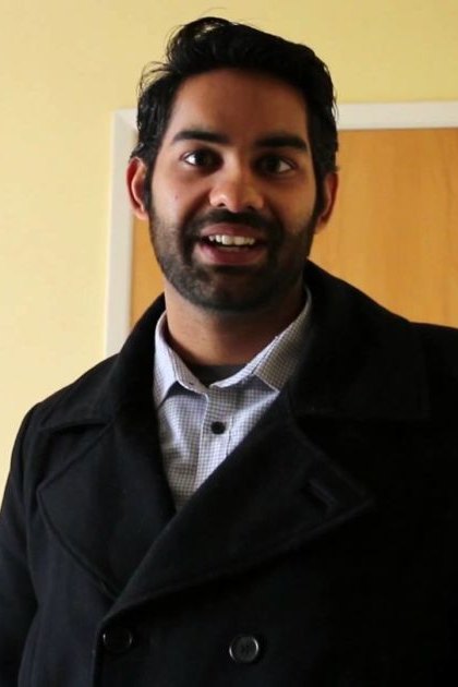 Young adult male wearing a dark overcoat with checkered shirt underneath in an indoor setting, with muted wall colors and furniture.