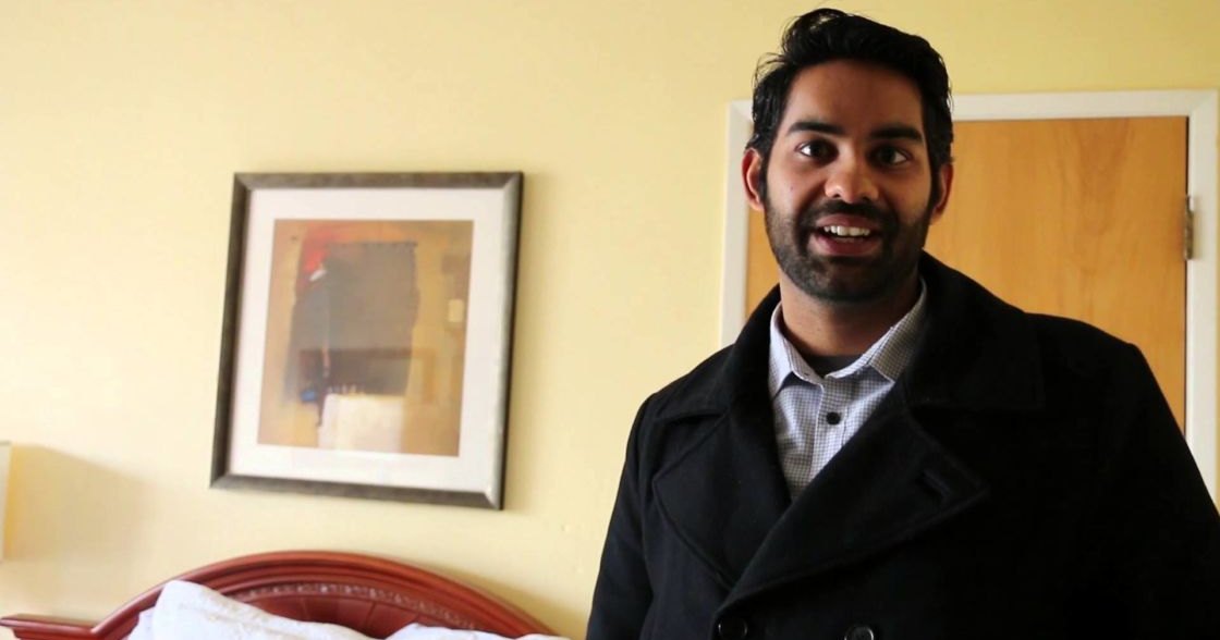 Young adult male wearing a dark overcoat with checkered shirt underneath in an indoor setting, with muted wall colors and furniture.
