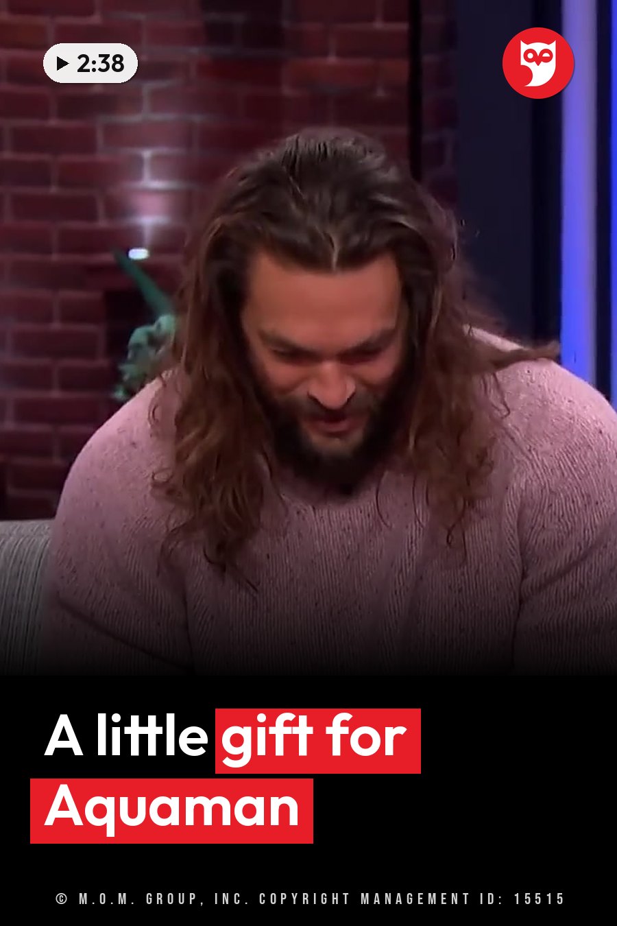 Jason Momoa, with long hair and a beard, wears a pink sweater and looks down at something in his lap. A headline runs in white letters across the lower third.
