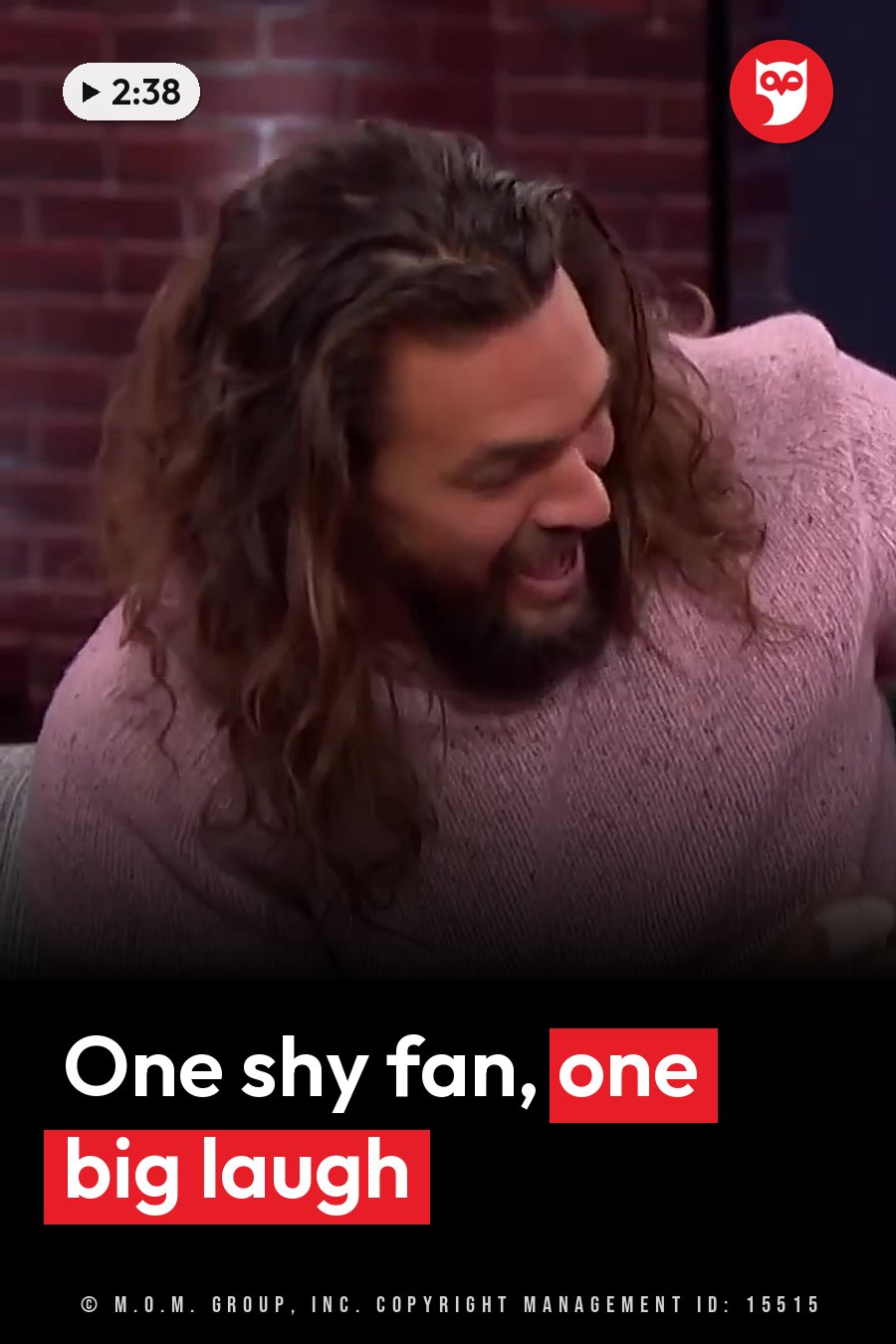 Jason Momoa, with long hair and a beard, laughs in a pink sweater in front of a brick wall. A headline runs in white letters across the lower third.