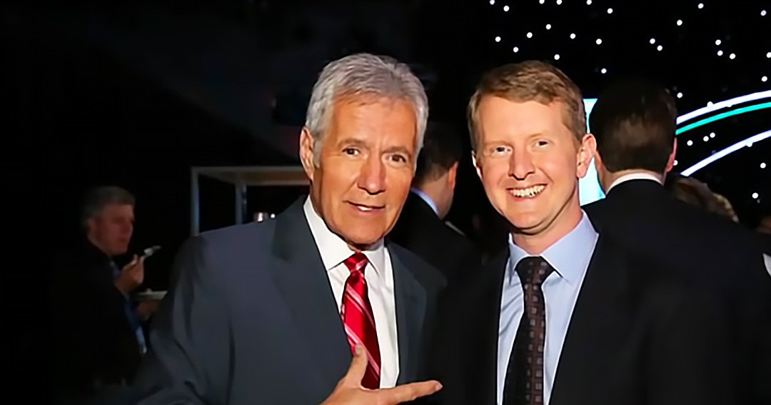 Two adult men in formal attire, one with gray hair and a red striped tie, another with brown hair and a patterned tie, standing indoors at an evening event space.