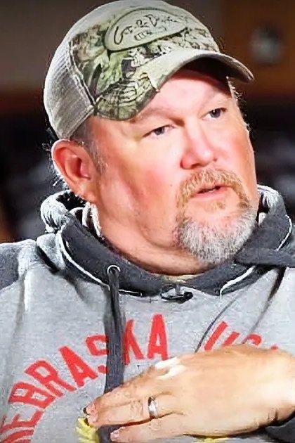 Older adult male wearing a gray hoodie with red lettering, camo baseball cap backward, and wedding ring in casual indoor setting