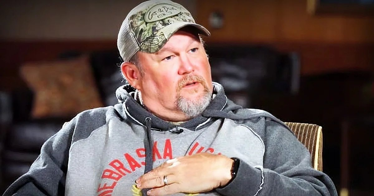 Older adult male wearing a gray hoodie with red lettering, camo baseball cap backward, and wedding ring in casual indoor setting