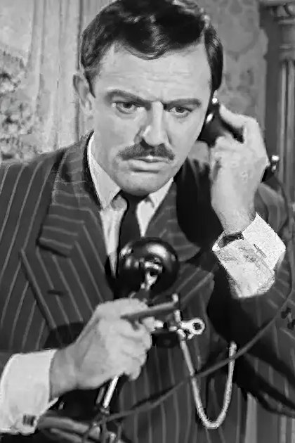 Adult male in a pinstriped suit jacket holding an old-fashioned telephone receiver, wearing gloves; adult female in dark-colored dress with fur collar or trim, arms crossed over chest. Ornate vase and intricate patterns suggest formal interior space from the mid-20th century.