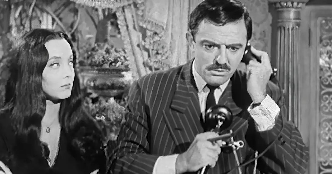 Adult male in a pinstriped suit jacket holding an old-fashioned telephone receiver, wearing gloves; adult female in dark-colored dress with fur collar or trim, arms crossed over chest. Ornate vase and intricate patterns suggest formal interior space from the mid-20th century.