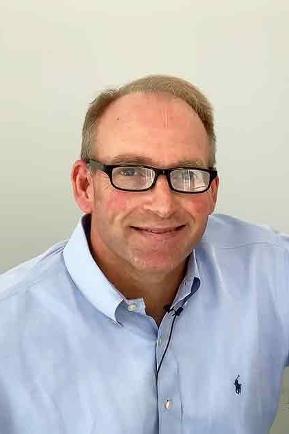 A middle-aged adult male wearing a light blue button-up shirt with black-rimmed glasses, indoors in an office or home environment.