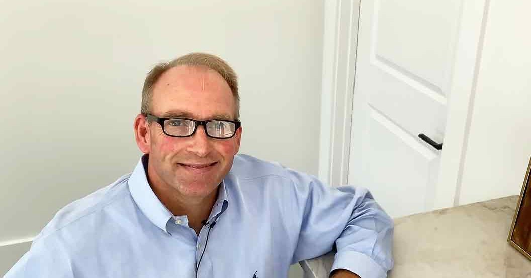 A middle-aged adult male wearing a light blue button-up shirt with black-rimmed glasses, indoors in an office or home environment.