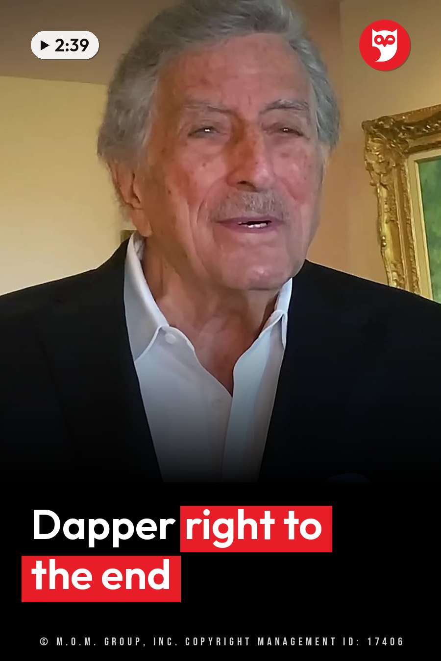 An older man with gray hair and a mustache, in a black jacket over a white shirt, speaks indoors near a gold-framed picture. A headline runs in white letters across the lower third.