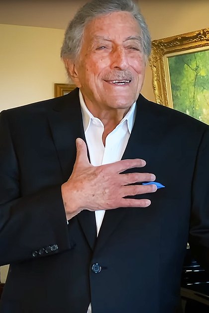 An older adult male wearing a black suit jacket with white shirt, standing indoors near a piano and framed photographs. The setting suggests a home office or living room with diffused natural light.