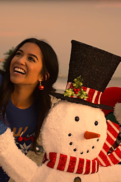 Beach scene at sunset with two young adults posing; one holds a snowman decoration and the other wears festive sweater and top hat, ocean waves visible in background