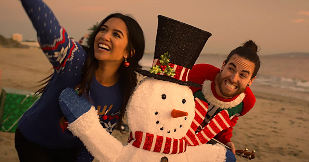 Beach scene at sunset with two young adults posing; one holds a snowman decoration and the other wears festive sweater and top hat, ocean waves visible in background