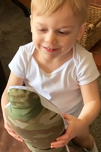 Indoors, young child wearing a white cotton T-shirt with 'I got my daddy back' text holding camouflage-patterned item, surrounded by adults dressed in winter and military attire, carpeted floor, artificial lighting, evening or night setting.