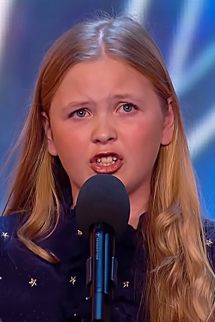A young girl in a loose-fitting, dark blue star-patterned hooded garment performs on stage at what appears to be a talent show under bright indoor lighting with a colorful backdrop.