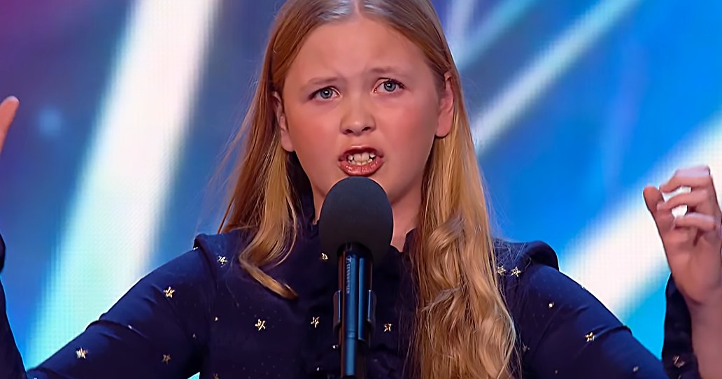 A young girl in a loose-fitting, dark blue star-patterned hooded garment performs on stage at what appears to be a talent show under bright indoor lighting with a colorful backdrop.