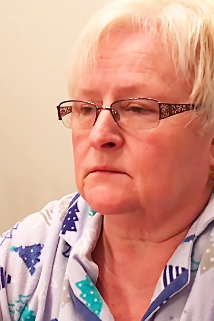 An older adult female in a home setting, wearing patterned Christmas-themed pajamas with long sleeves. The scene is indoors, possibly during winter.