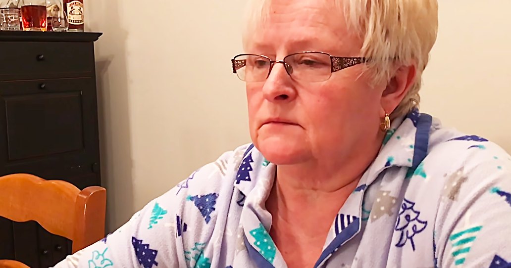 An older adult female in a home setting, wearing patterned Christmas-themed pajamas with long sleeves. The scene is indoors, possibly during winter.
