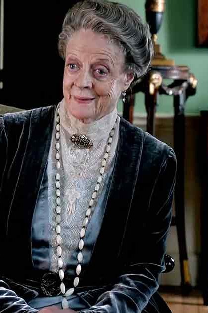 An older adult woman seated in a formal setting, wearing a dark velvet jacket with intricate embroidery and a light-colored lace blouse, accessorized with a long pearl necklace and earrings; partially visible adult male in a dark coat or jacket and possibly a top hat. Ornate furniture and historical decor suggest an indoor Victorian-era scene.