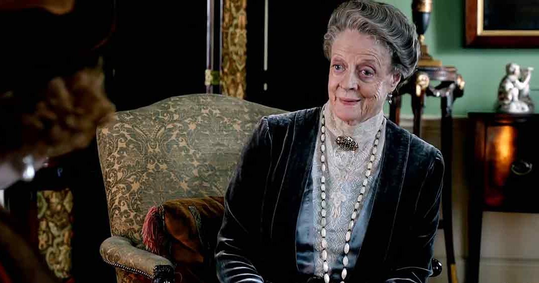 An older adult woman seated in a formal setting, wearing a dark velvet jacket with intricate embroidery and a light-colored lace blouse, accessorized with a long pearl necklace and earrings; partially visible adult male in a dark coat or jacket and possibly a top hat. Ornate furniture and historical decor suggest an indoor Victorian-era scene.