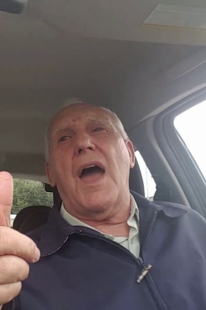 An older adult male passenger in a dark blue zip-up jacket sits beside a middle-aged man wearing a white t-shirt with a gold chain necklace, inside a car on an outdoor road with green trees visible through the windows.