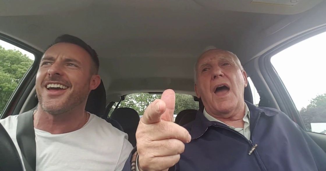 An older adult male passenger in a dark blue zip-up jacket sits beside a middle-aged man wearing a white t-shirt with a gold chain necklace, inside a car on an outdoor road with green trees visible through the windows.
