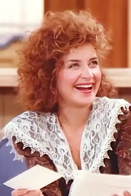 A middle-aged adult female wearing a white lace top with brown patterns and long sleeves, standing in an indoor kitchen or café setting with bright, natural lighting.